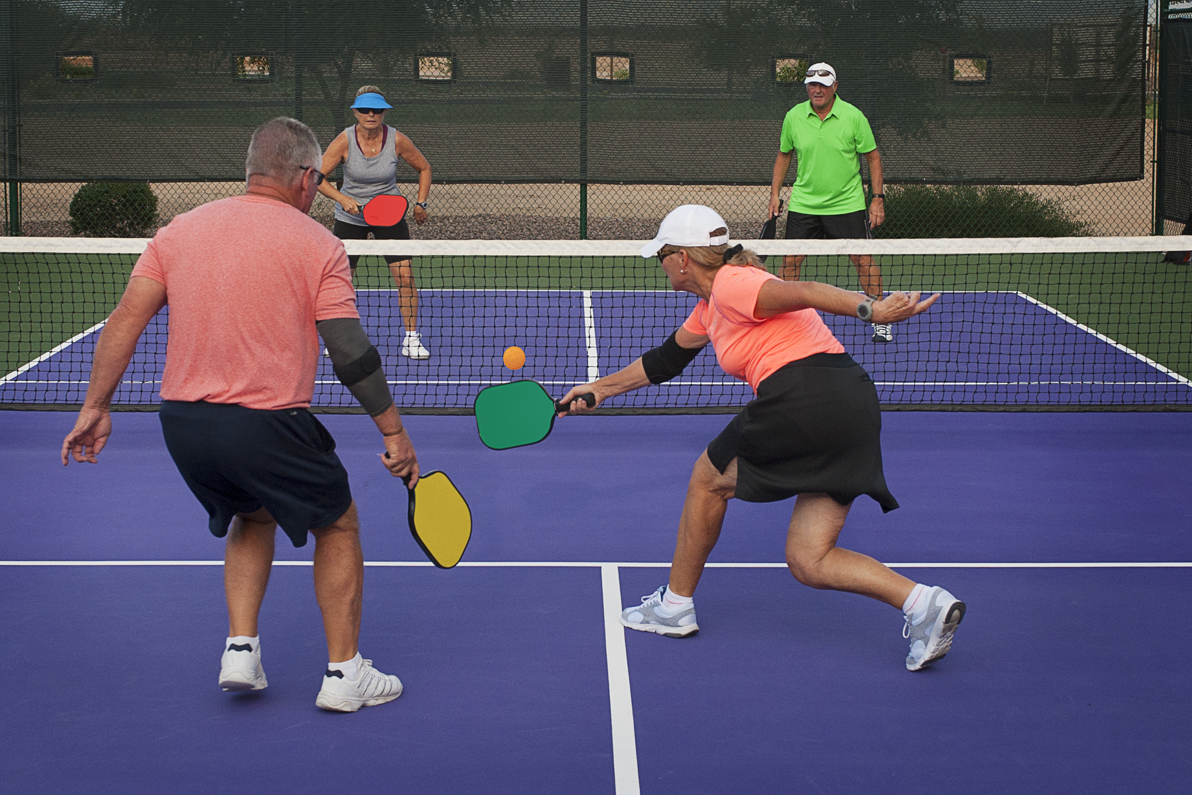 Four people playing pickleball on a purple court; a woman in a black skirt lunges to hit the yellow ball with a green paddle.